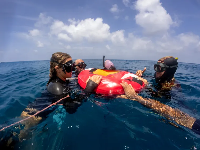Three freedivers at the surface around the buoy, in masks and wetsuits.