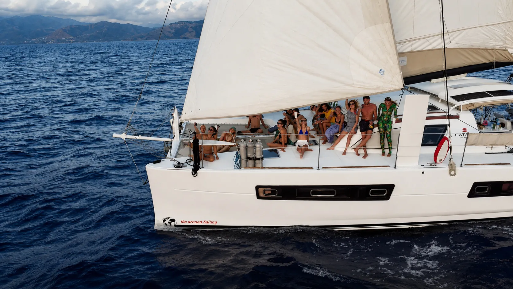 Guests sitting on the bow of IKIGAI under way, the coast behind them