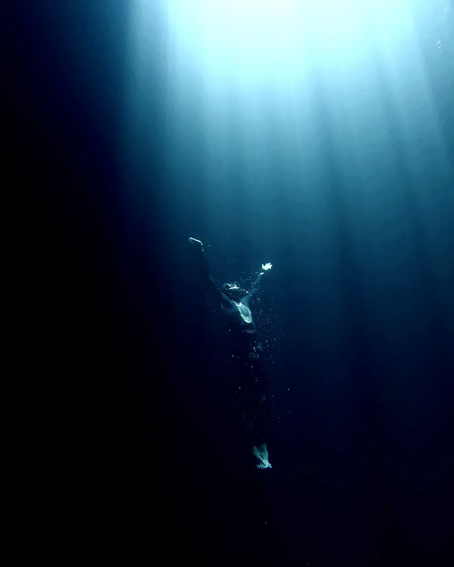 A freediver ascending through a shaft of light in the deep blue.